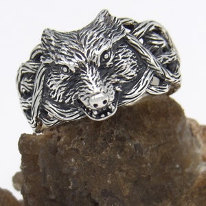 May include: A silver ring with a wolf head design. The wolf's head is detailed and realistic, with sharp teeth and a fierce expression. The ring is set on a rough, brown rock.