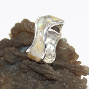 May include: A silver ring with an abstract design. The ring features a central section with a mosaic of light-colored stones, possibly opal, set within a polished silver band. The ring is displayed on a textured, dark brown surface.