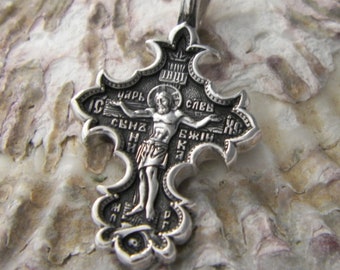 May include: A silver pendant in the shape of a cross with intricate details and a figure of Jesus Christ on the front. The cross is decorated with Cyrillic lettering.