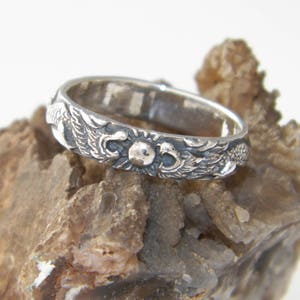 May include: A silver ring with a detailed design of two birds flanking a sun with a raised center. The ring is resting on a rough, brown rock.