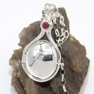 Puede incluir: Un collar de plata con un medallón que tiene un acento de piedra preciosa roja. El medallón es redondo y tiene una tapa con bisagras. La cadena es de plata y tiene un cierre de langosta.