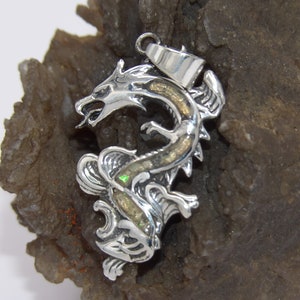 Könnte beinhalten: Silberner Drachenanhänger mit schillerndem Opal-Inlay. Der Drache ist in einer defensiven Haltung mit ausgebreiteten Flügeln zusammengerollt. Der Anhänger befindet sich auf einem grauen Hintergrund.