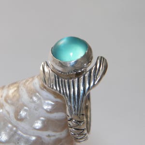 May include: A silver ring featuring a turquoise blue cabochon gemstone set in a textured silver band. The band is shaped like a mermaid's tail.