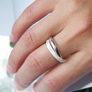 May include: A silver ring with a textured band. The ring is worn on a finger.