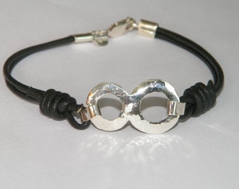 Op de afbeelding: Een zwart leren armband met een zilveren oneindigheidssymbool bedel. De bedel heeft een gehamerde textuur en bestaat uit twee in elkaar grijpende cirkels.