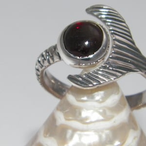 May include: A silver ring with a dark red gemstone set in a round bezel. The ring band is textured and the setting is shaped like a stylized fish tail.
