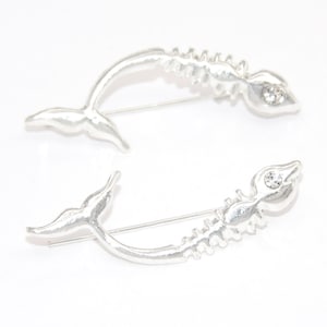 May include: Two silver fish skeleton earrings with a single rhinestone on each. The earrings are curved and have a tail fin.