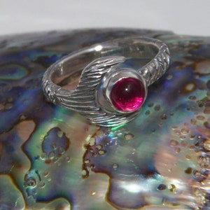 May include: A silver ring featuring a pink gemstone set within a textured, feather-like design.