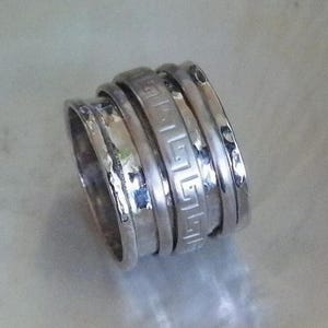 May include: A silver ring with four bands, one of which has a Greek key design. The bands are stacked on top of each other and have a hammered texture.