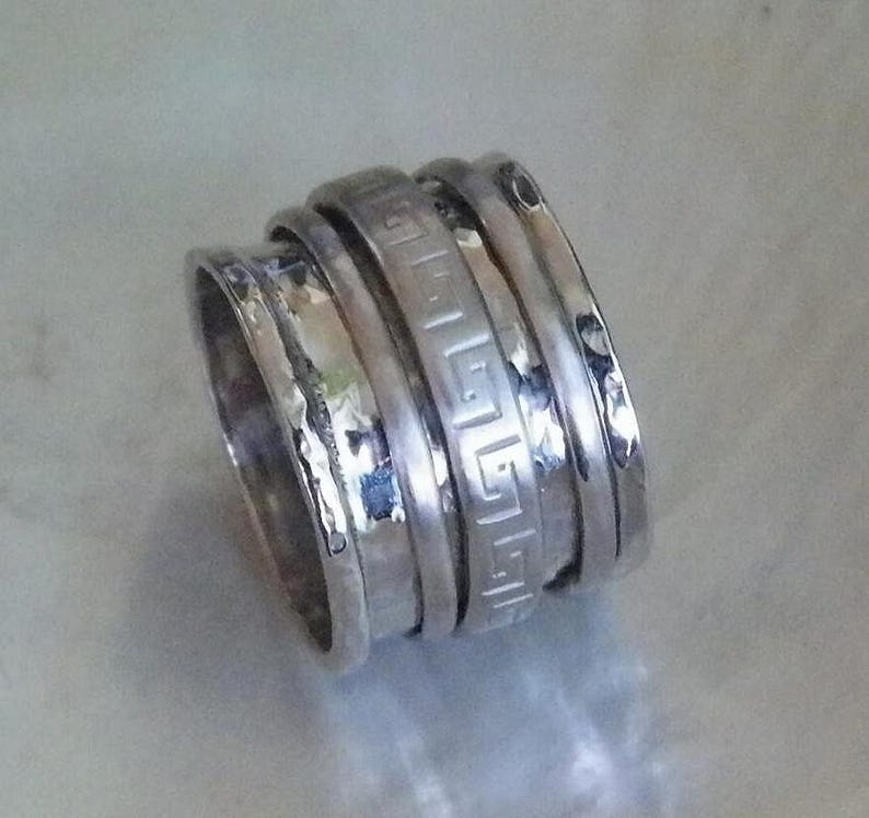 May include: A silver ring with four bands, one of which has a Greek key design. The bands are stacked on top of each other and have a hammered texture.