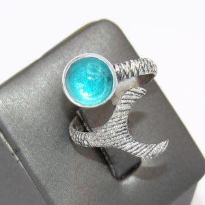 May include: A silver ring with a turquoise blue glass stone set in the center. The ring is shaped like a mermaid tail.