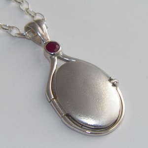 Puede incluir: Collar con dije ovalado de plata con un acento de piedra preciosa roja. El dije tiene una abertura con bisagra y un acabado liso y pulido.
