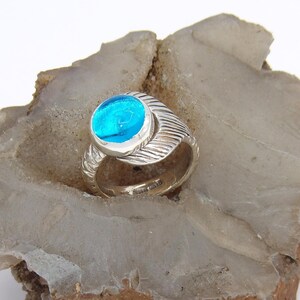 May include: A silver ring with a round, bright blue stone. The ring has a textured band with a swirling design. The ring is set on a rough, light brown and gray stone.