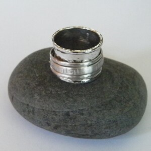 K&ouml;nnte beinhalten: Ein silberner Ring mit geh&auml;mmerter Oberfl&auml;che und einem griechischen Schl&uuml;sselmuster. Der Ring liegt auf einem grauen Stein.