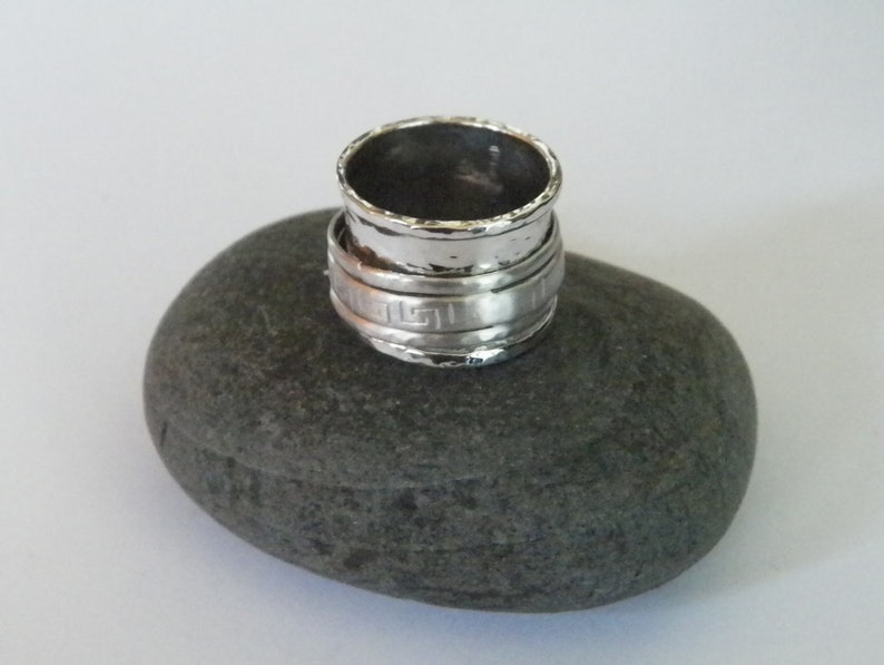 K&ouml;nnte beinhalten: Ein silberner Ring mit geh&auml;mmerter Oberfl&auml;che und einem griechischen Schl&uuml;sselmuster. Der Ring liegt auf einem grauen Stein.