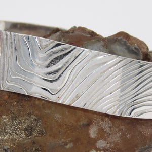 May include: A silver bangle bracelet with a wavy, textured design. The bracelet is made of polished metal and has a smooth, rounded surface.