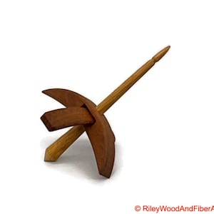 May include: A wooden drop spindle for hand spinning yarn. The spindle has a shaft and a whorl, with a natural wood finish. The whorl has a distinctive shape, and the spindle is set against a white background.