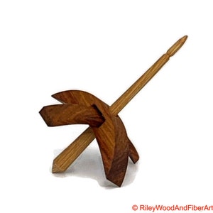 May include: A wooden drop spindle with a dark brown whorl and a light brown shaft. The whorl has a geometric design with three angled blades. The spindle is used for hand spinning fibers into yarn.