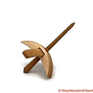 May include: A wooden drop spindle, a tool for hand spinning yarn. The spindle features a pointed shaft and a wooden whorl. The whorl has a crescent shape and is made of light-colored wood. The shaft is a darker shade of brown.
