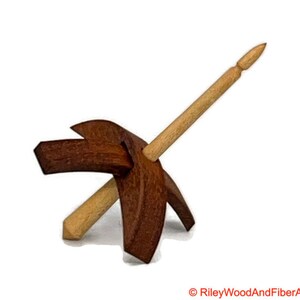 May include: A wooden drop spindle with a dark brown whorl and a light brown shaft. The spindle is designed for hand spinning yarn. The whorl has a unique shape with angled arms. The shaft is inserted through the center of the whorl.