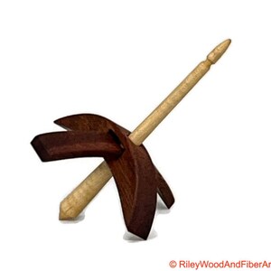 May include: A wooden spinning wheel with a brown base and a light wood spindle. The spindle is attached to the base with a metal pin.