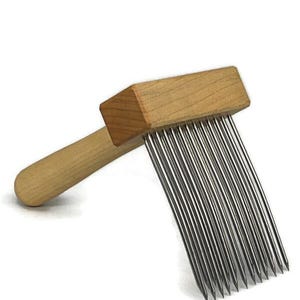 May include: A wooden fiber art tool with a rectangular head and a rounded handle. The head has a row of closely spaced, thin, metal tines. The tool is made of light-coloured wood and metal.