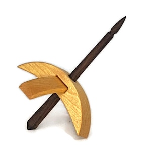 May include: A wooden drop spindle with a dark brown shaft and a light brown, crescent-shaped whorl. The whorl has a smaller, rectangular piece attached. The spindle is designed for spinning fibers into yarn.