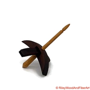 May include: A wooden drop spindle with a dark brown whorl and a light brown shaft. The spindle is designed for hand spinning fibers into yarn. The whorl has a unique shape with a geometric design.