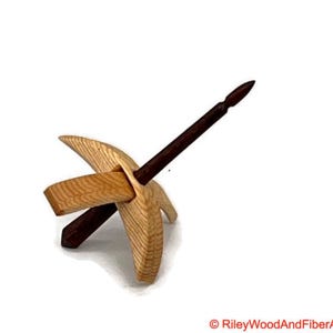 May include: A wooden drop spindle with a natural wood finish. The spindle has a dark brown shaft and a light-coloured wooden whorl. The whorl has a unique shape with curved edges and a rectangular crossbar. The spindle is designed for spinning yarn.