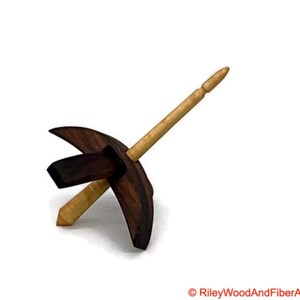 May include: A wooden drop spindle with a dark brown whorl and a light brown shaft. The whorl has a unique shape with curved edges and a rectangular center. The shaft is tapered at both ends, designed for spinning yarn.