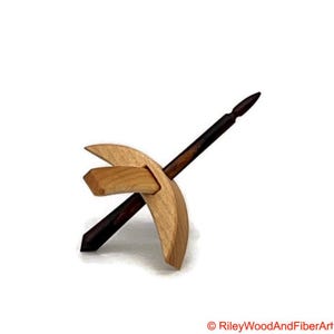 May include: A wooden drop spindle featuring a light-coloured wooden whorl and a dark-coloured shaft. The whorl has a distinctive curved design, and the shaft tapers to a point. The spindle is designed for spinning fibre into yarn.