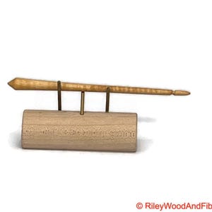 May include: A light-colored wooden spindle, supported by brass-colored metal rods and a cylindrical wooden base. The spindle tapers to a point on one end and has a rounded tip on the other. The base has etched text.