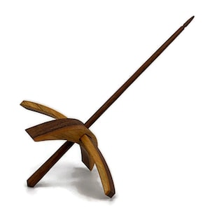 May include: A wooden drop spindle with a dark brown shaft and a cross-shaped whorl. The whorl is made of lighter-colored wood and has curved arms. The spindle is designed for spinning yarn and fiber.
