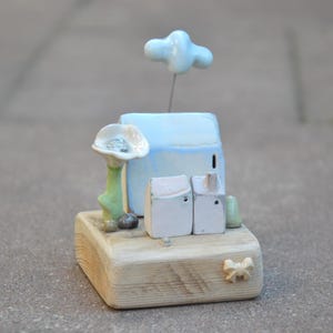 May include: A ceramic sculpture on a wooden base featuring a large light blue house, two smaller white houses, a green-stemmed white flower, and a light blue cloud on a wire. A small beige bow is attached to the front of the wooden block base.