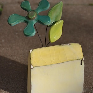Puede incluir: Un recipiente de pared de cerámica con forma de casa en amarillo pálido con un acabado de esmalte agrietado en el techo. Un tallo de alambre decorativo presenta una flor verde azulado de cinco pétalos y dos hojas verde brillante.