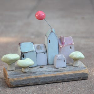 Op de afbeelding: Een keramisch miniatuurdorp met vier kleine huisjes in lichtblauw, wit en roze, geplaatst op een rechthoekig stuk verweerd hout. Twee lichtgroene paddenstoelfiguren flankeren de huisjes, samen met een roze ballon aan een ijzerdraadje.