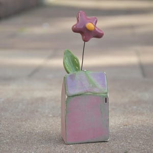 Könnte beinhalten: Eine kleine Keramikhausfigur mit glänzender rosa und grüner Oberfläche, die mit einem grünen Blatt und einer violetten Blume mit gelbem Zentrum versehen ist, welche durch einen dünnen Metalldraht am Dach befestigt sind als Tischschmuck.