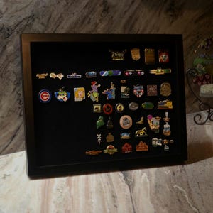 May include: A black rectangular shadow box display case containing a collection of various small metal enamel pins arranged on a black velvet backing. The pins feature diverse shapes, colors, and graphic designs, including logos and illustrations.