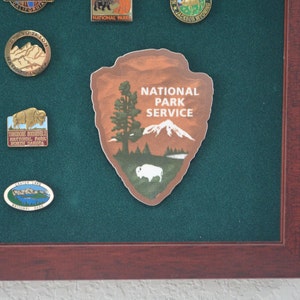 May include: A brown and white National Park Service patch with a mountain, a tree, and a bison. The text "National Park Service" is printed in white on the patch.