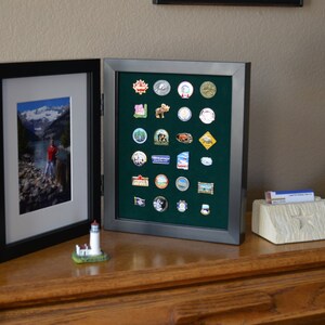 May include: A black framed shadow box with a green felt interior displaying a collection of colorful enamel pins. The shadow box is on a wooden surface next to a framed photograph and a small lighthouse figurine.