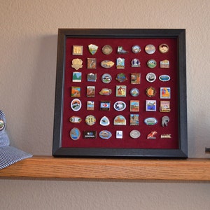 May include: A black framed shadow box with a red velvet background displaying a collection of colourful enamel pins. The pins feature various national park logos and designs.