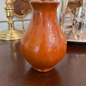 May include: A small, orange-brown ceramic vase with a rounded body and a slightly flared rim. The vase has a glossy finish and sits on a small, circular base. The background includes a gold-framed mirror and silver serving pieces.