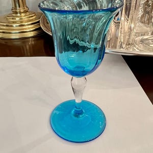 May include: A vibrant, turquoise-colored glass goblet with a clear stem and base. The bowl has a textured, swirling design. The goblet is likely used for serving beverages and adds a pop of color to any table setting.