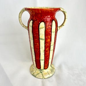 May include: A decorative ceramic vase with a red background and vertical white stripes. The vase has two handles and a patterned base. The vase is approximately 10 inches tall.