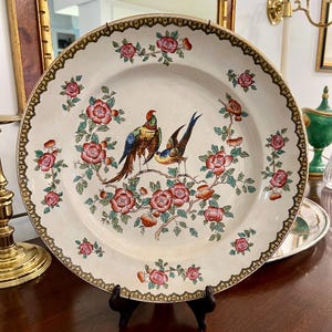 May include: A large, antique decorative plate with a cream-colored background. The plate features a detailed design of two birds perched on a flowering branch with pink blossoms and green leaves. The rim has a brown and gold patterned border.
