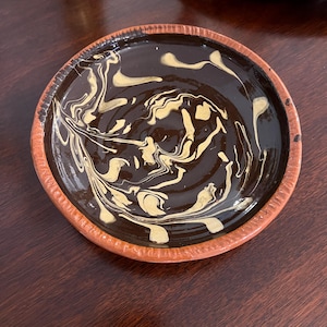 May include: A round ceramic dish with a terracotta-colored rim and a dark brown interior glaze. The center features a swirling, marbled pattern of light gold paint that creates organic, flowing lines across the dark surface.