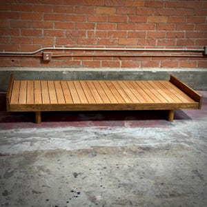 May include: A wooden daybed with a slatted top and a low profile. The bed is made of light brown wood and has a simple, minimalist design. The bed is approximately 78 inches long and 36 inches wide.