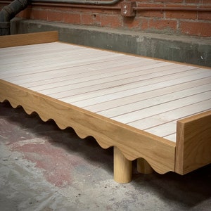 May include: A light-colored wooden bed frame with a scalloped edge design. The bed has a slatted base and rounded legs. The frame is made of light-colored wood, and the design is modern and minimalist.