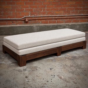 May include: A rectangular daybed featuring a dark brown wooden frame with a low-profile base and four sturdy legs. The top consists of a thick, cream-colored textured fabric cushion that spans the entire length of the wooden platform base.