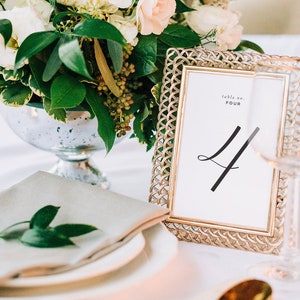 Peut inclure: Un cadre de num&eacute;ro de table dor&eacute; avec une carte blanche sur laquelle est inscrit "table no. FOUR" en noir. Le chiffre quatre est &eacute;crit en cursive. Le cadre est pos&eacute; sur une nappe blanche avec une serviette verte et un vase de fleurs blanches.